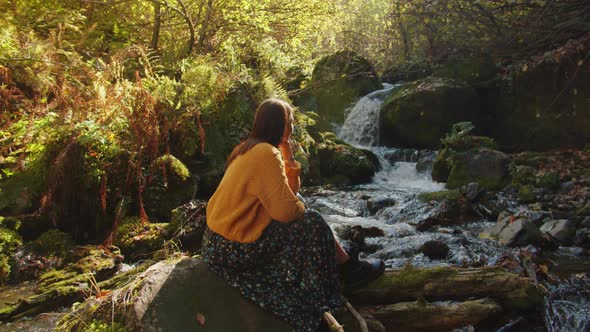 Young Woman is Relaxing Nearby the Flowing Mountain Stream Among the Scenic Autumn Forest