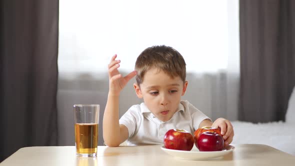 Happy Child Is Drinking Apple Juice and Eating an Apple, Sitting at a Table in the Room. Children's alt
