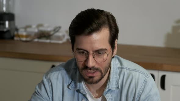 Close-Up of a Young European Man Talking to a Colleague, Working Online at Home in the Kitchen alt