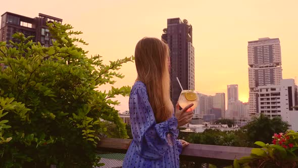 Young Woman in Gown Holding Cocktail on Terrace of Rooftop Bar During Sunset alt