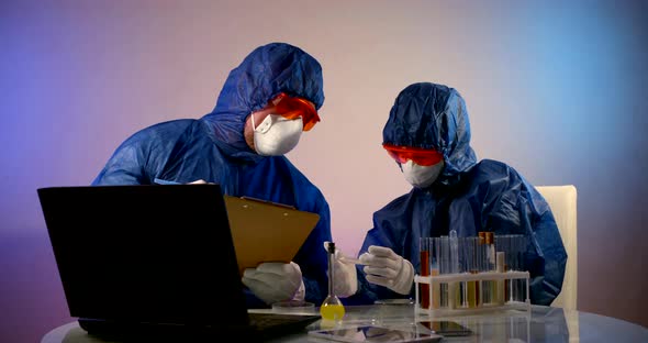 People in Protective Clothing with Hoods, Respirators, Glasses and Gloves Are Sitting at a Table alt