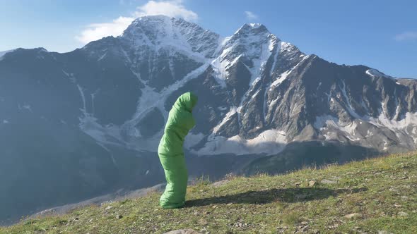 Funny Male Traveler Jumps in a Sleeping Bag Against the Backdrop of High Snowy Mountains in Slow alt