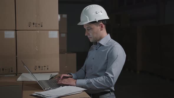 Man in a Warehouse Works with Laptop on Background of Cardboard Boxes