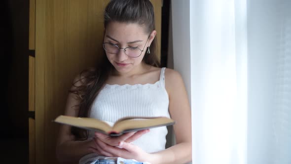 girl with glasses reads a book near the window and drinks tea or coffee alt