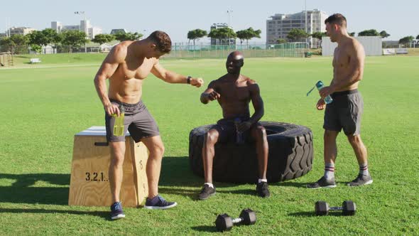 Three fit shirtless diverse men resting, drinking water and talking after exercising outdoors alt