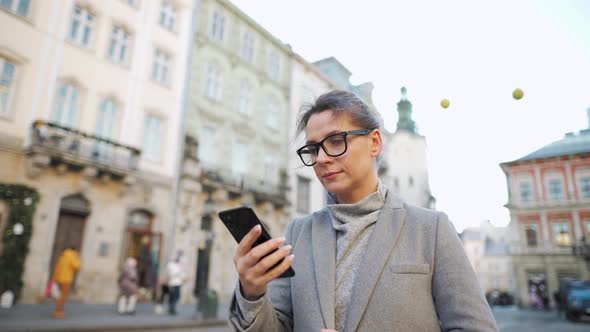 Woman with Glasses Wearing a Coat Walking Down an Old Street and Using Smartphone alt
