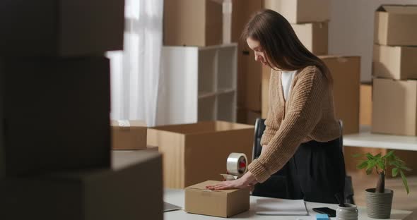 Female Worker of Small Domestic Warehouse Is Using Rollon Tape Dispenser for Packing Box with Goods alt