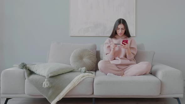 Young woman with glasses sitting on couch and making online purchases with credit card.