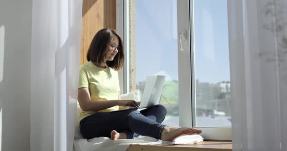 Young Beautiful Woman is Sitting on the Windowsill and Chatting Using a Laptop