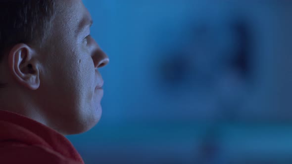 A Man Sits at the TV at Night Watching a Movie Multicolored Reflections on His Face Closeup