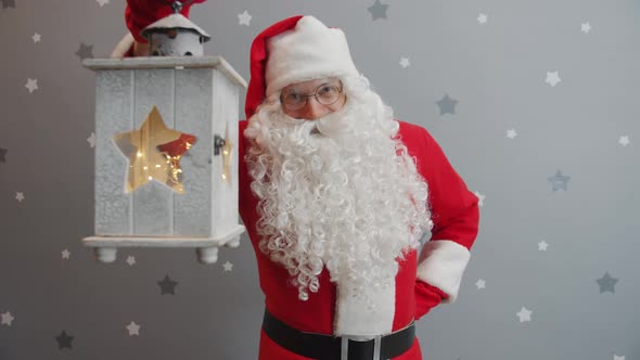 Slow Motion Portrait of Smiling Santa Holding Lantern and Looking at Camera on Glittery Background alt