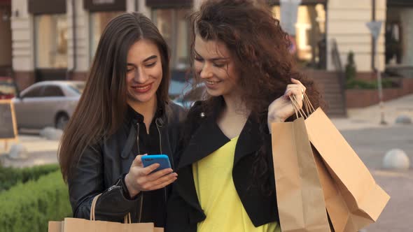 Girl Shows Her Friend Something on Her Smartphone on the Street of the City alt