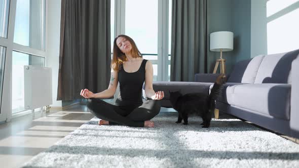Young Caucasian Woman in Black Jumpsuit Sitting in Lotus Position and Meditating Yoga at Home alt