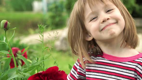 Little happy girl in summer park smells bud of purple peony flower, looks at camera and smiles.