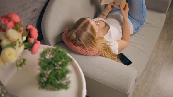 Young Woman Reading Book Lying on Sofa with Mobile Phone Next to Her alt