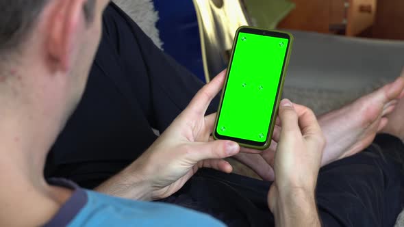 A Man on the Couch With a Phone in His Hands With a Green Screen Back View alt
