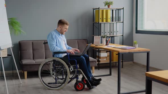 Disabled Person Freelancer is Working with Laptop Typing Sitting in Wheelchair at Office alt
