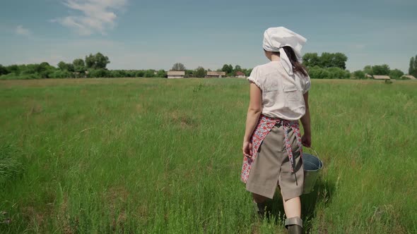 Young Milkmaid in Sunny Summer Carries a Bucket and Walks on the Grass alt
