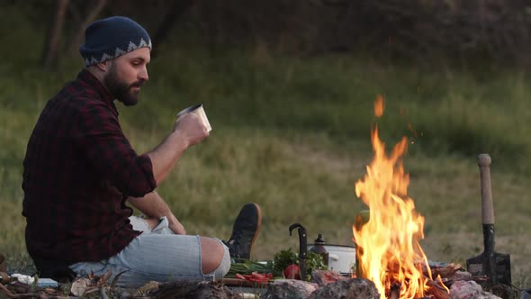 Tourist Hiker Sits By Campfire Looks Into Blazing Flame and Drinks Hot Tea Coffee From Metal Cup