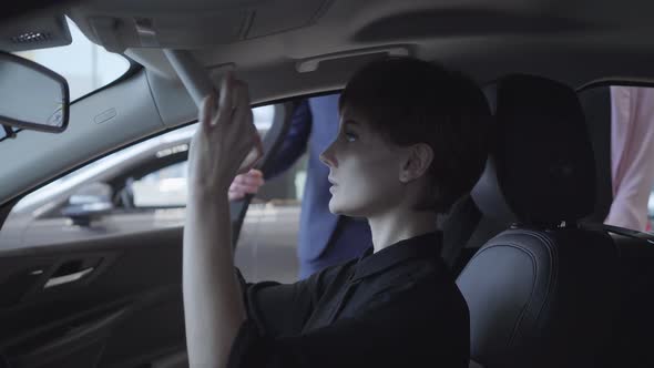 Young Woman Examines New Auto While Sitting in Passenger Compartment of the New Car in Car Showroom alt