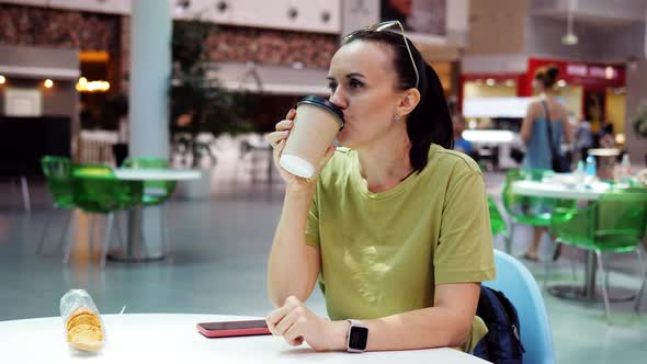 A Woman Drinks Coffee From a Cup in a Food Court Sitting at a Table alt
