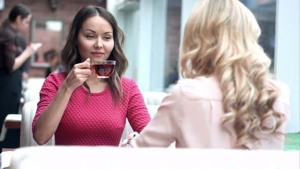 Two Young Beautiful Girls Sitting in Urban Cafe with Coffee and Talking alt