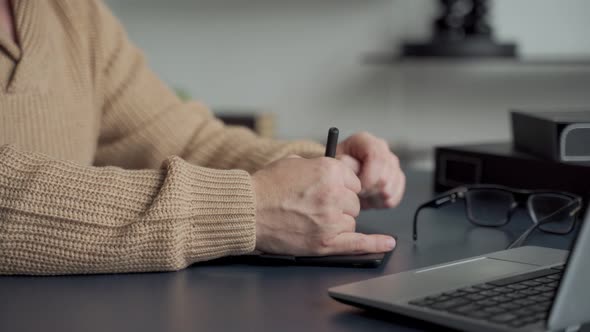 An Elderly Man with a Gray Beard Draws with a Pen on a Tablet at Home in the Living Room alt