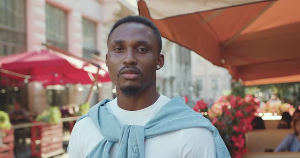 African American Standing on the Background of Bright Cozy Street Cafe and Looking at Camera alt