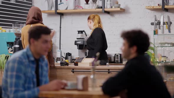 Young Slim Muslim Woman Receiving Takeaway Order in Cafe with Blurred Men Talking at Front alt