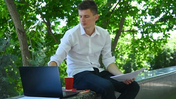Young Businessman Sitting In Park And Examines Business Report
