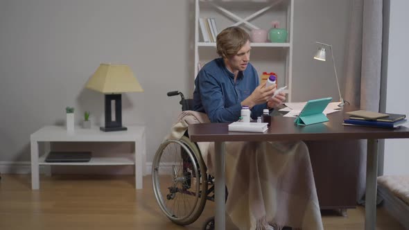 Wide Shot Young Paraplegic Man in Wheelchair Consulting Doctor Online Talking Showing Pills at alt