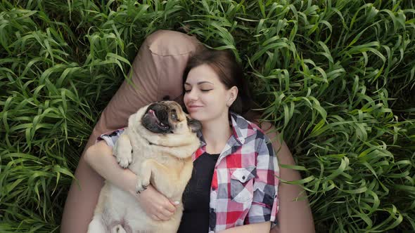 Woman Lies with a Pug in the Field on Bag Chair Kiss and Hugs Dog Playing in Nature alt