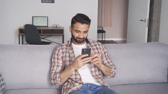 Young Attractive Indian Man Sitting on Sofa Holding Cellphone at Home alt