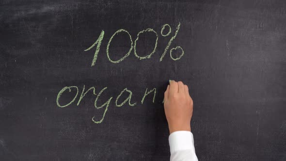 A man's hand writes the phrase 100% organic in green chalk on a chalkboard alt