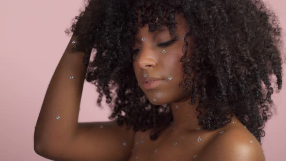Mixed Race Black Woman with Curly Hair Covered By Crystal Makeup on Pink Background in Studio alt