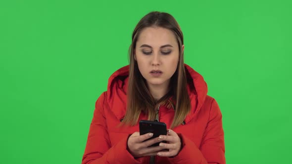 Portrait of Lovely Girl in a Red Down Jacket Is Texting on Her Phone. Green Screen alt