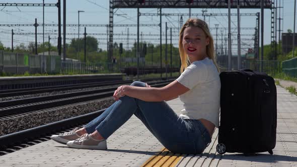 A Young Beautiful Woman Sits Next To a Suitcase on a Train Station Platform and Smiles at the Camera alt