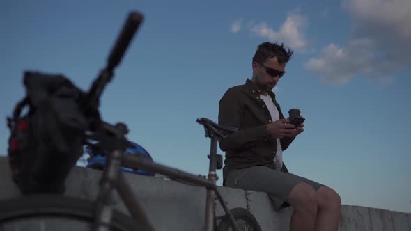 Man Cyclist Resting Overlooking Sea Drinking Coffee and Using Smartphone While Sitting on Embankment alt