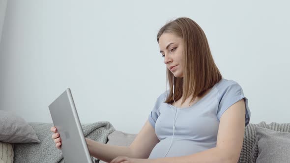 A Pregnant Woman in Home Clothes is Sitting on a Sofa and Using a Laptop alt