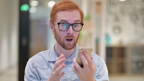 Portrait of Casual Redhead Man Celebrating Success on Smartphone  alt
