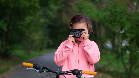 A little girl in a pink medical mask takes photos with an old film camera in a Park near a Bicycle.
