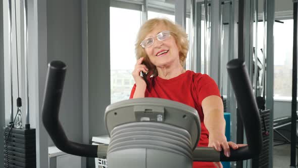 Smiling Elderly Woman Engaged in Cardio Training on Exercise Bike Talking on Phone alt