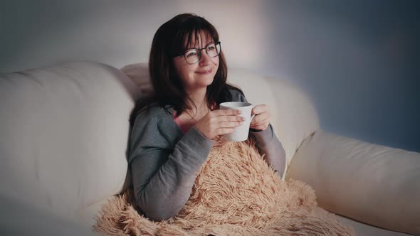 Happy Woman with a Glass of Coffee on the Sofa at Night in Front of the TV Switches Channel Enjoy