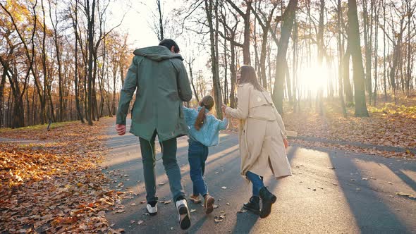 Young Parents Holding Hands of Little Daughter Laughing Playing Walking in Autumn Park alt