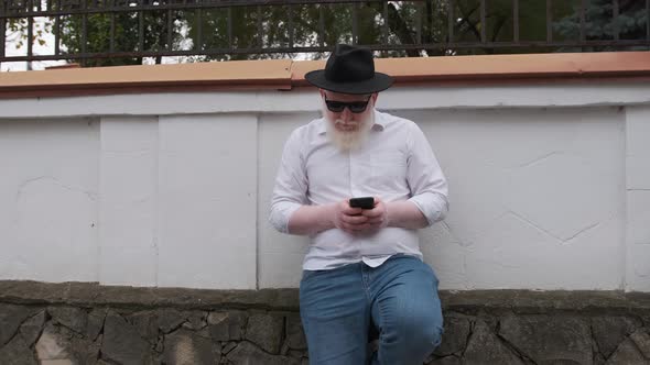 Old Man with Albinism Browsing on the Internet While Sitting Next To His House alt