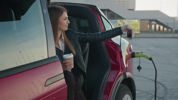 Charming Woman Sitting Inside Charging Electric Car with Modern Smartphone and alt