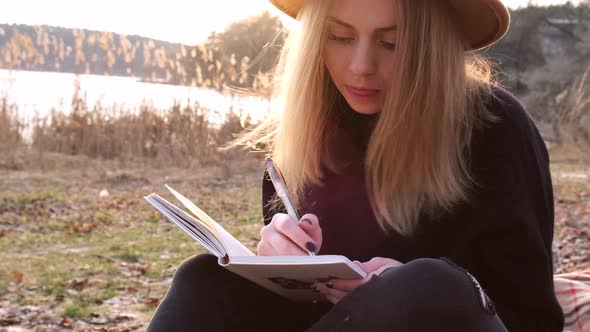 Caucasian Blonde Woman with Beige Hat in Black Sweater Write on Notebook in the Countryside alt