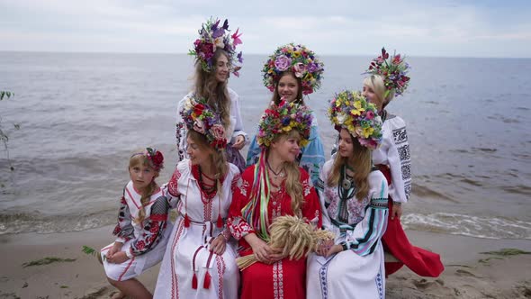 Group of Ukrainian Young Women and Girl Sitting on Sandy River Beach Smiling Looking Around alt