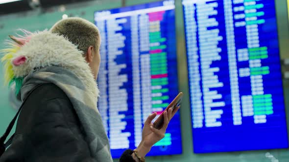 Young Girl with Short Hair Uses a Smartphone Looks Schedule on Scoreboard Airport and Checks Her