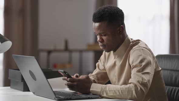 Black Male Office Worker or Freelancer is Using Smartphone During Work with Laptop Sitting at Table alt